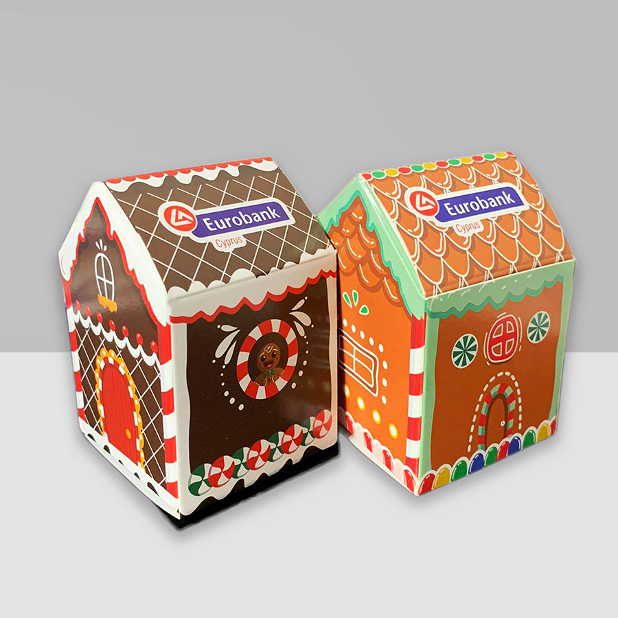 two cardboard ginger bread houses. Left one is brown, white and red, right one is orange, red and green. Both feature the Eurobank Cyprus logo.