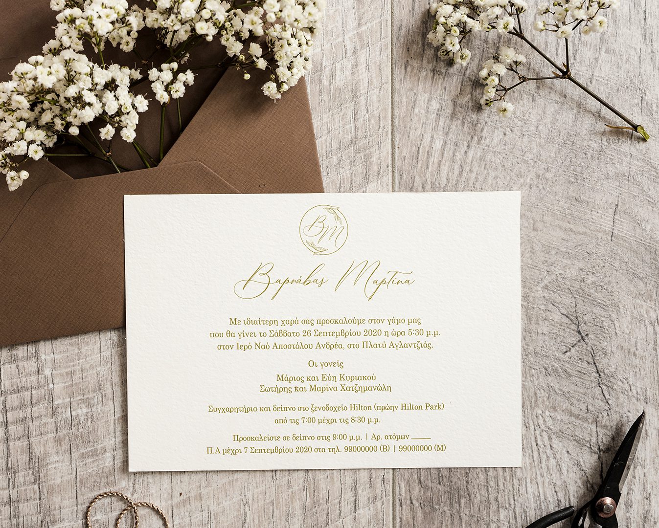 Wedding invitation printed on a textured cream color paper with gold test. Set on a wooden surface over a brown envelope with white flowers and two rings.