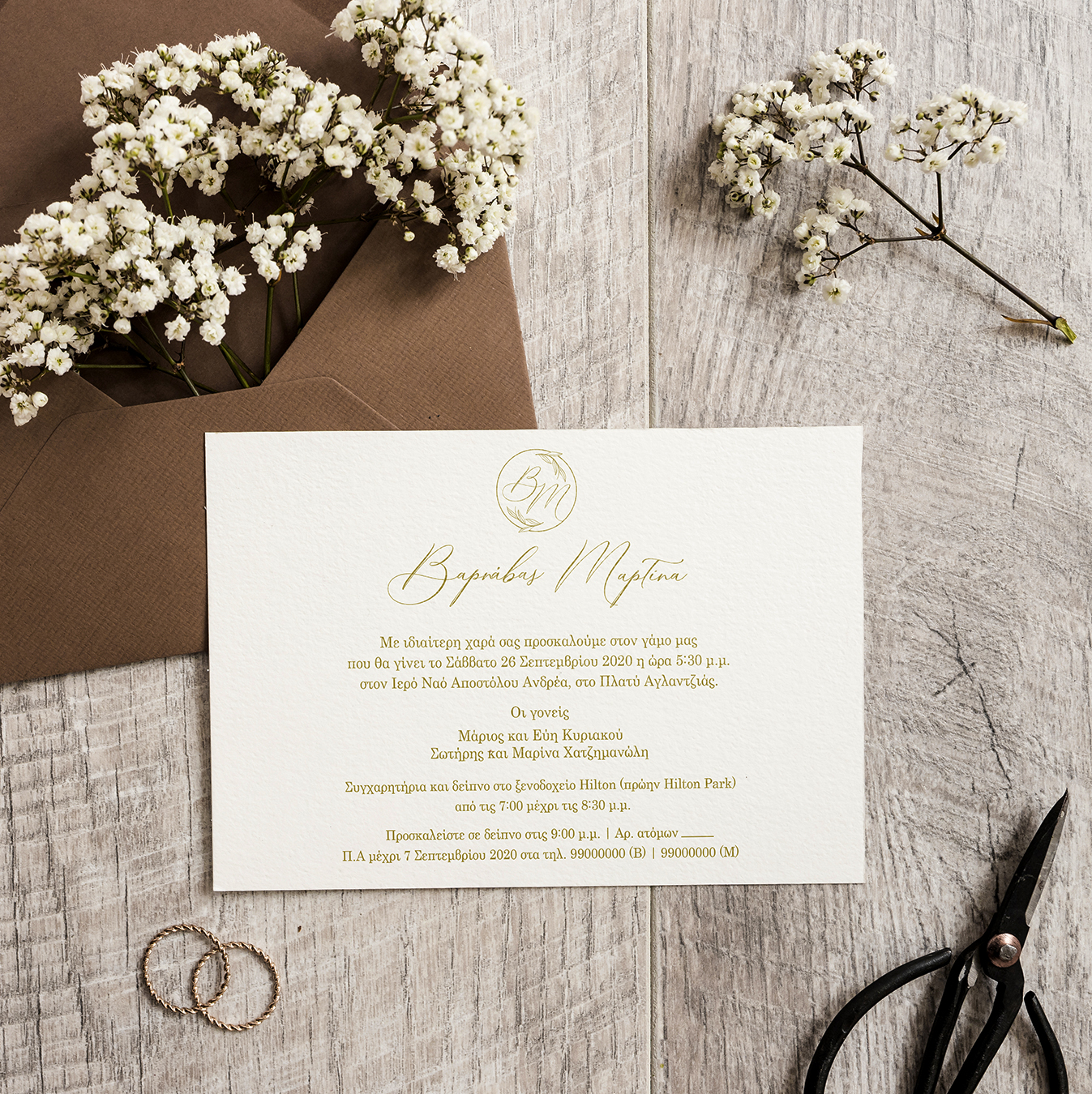 Wedding invitation printed on a textured cream color paper with gold test. Set on a wooden surface over a brown envelope with white flowers and two rings.